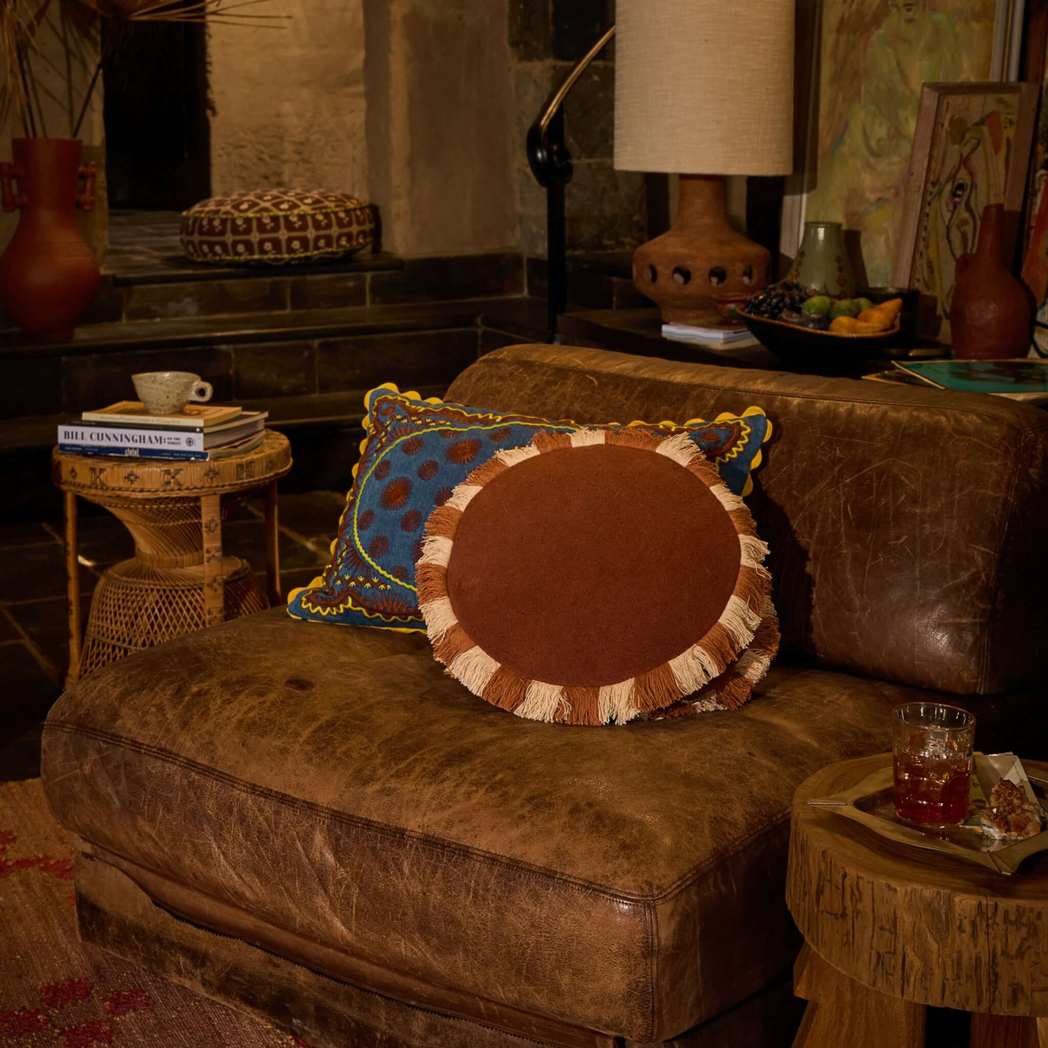 Sage x Clare Reese Round Cushion - Umber displayed on a plush leather sofa, featuring textured umber fabric and playful fringe.