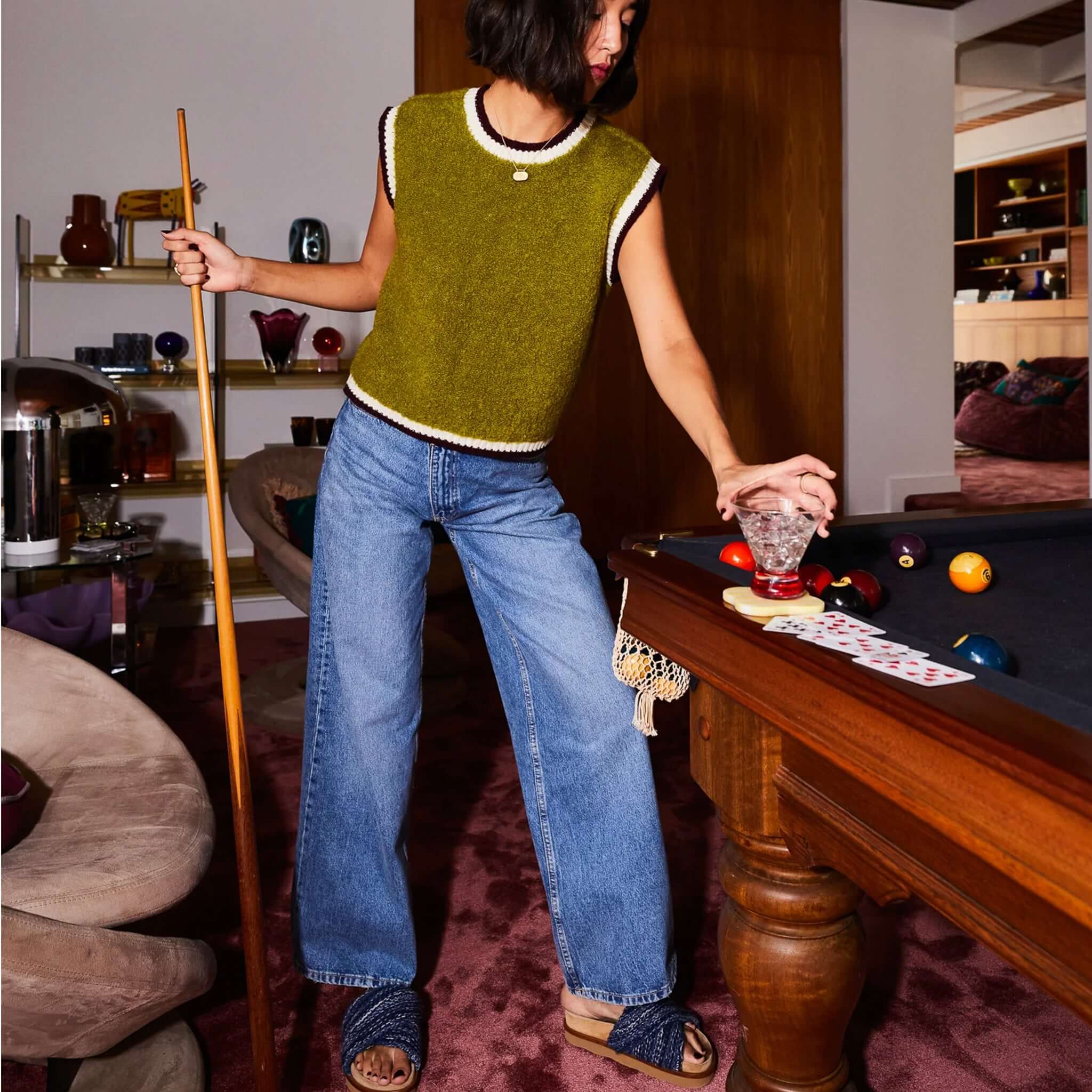 A woman wearing a green sleeveless top and wide jeans is playing pool, holding a glass in a stylish room.