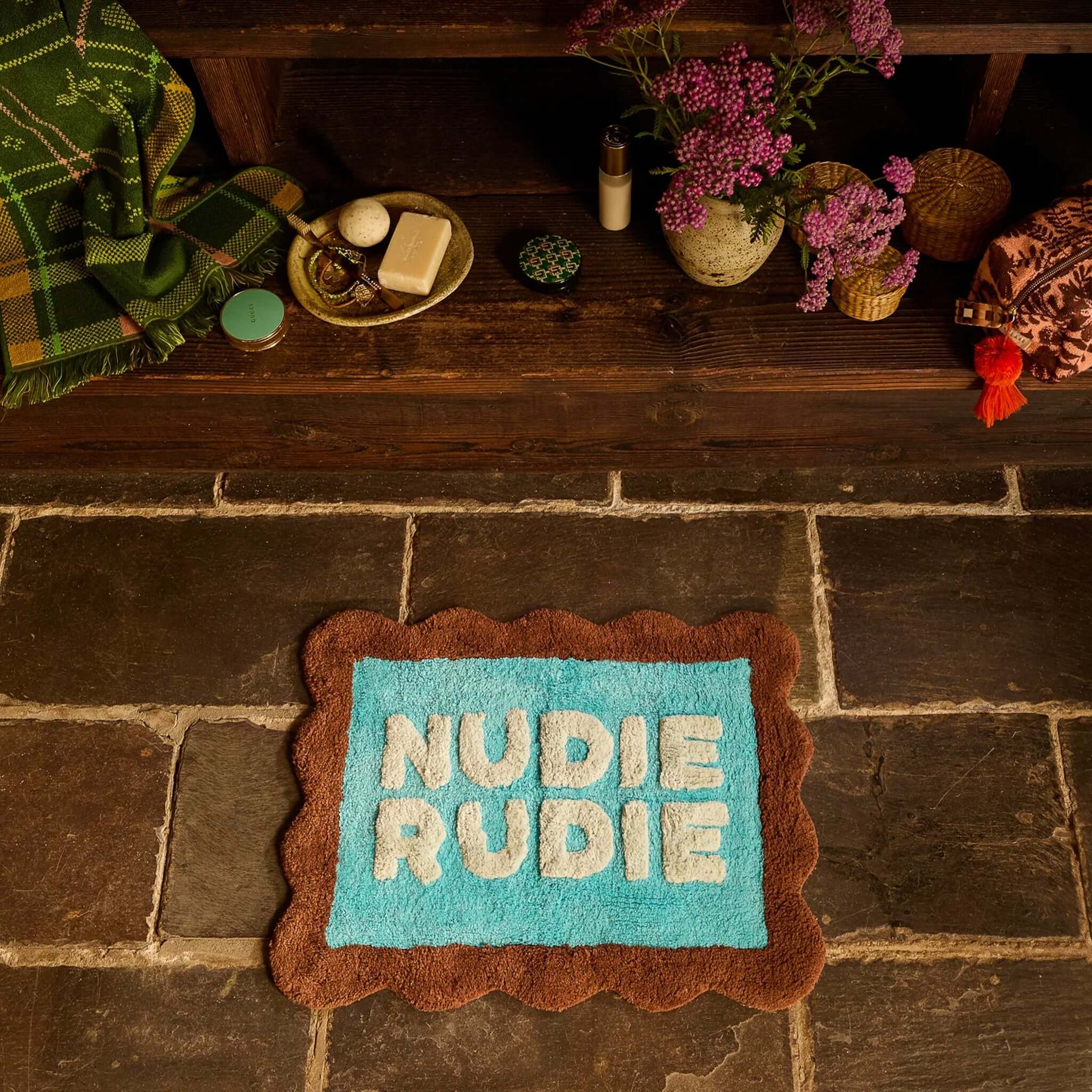 Sage x Clare Romy Nudie Bath Mat Mini - Turquoise on stone floor with flowers and decor.