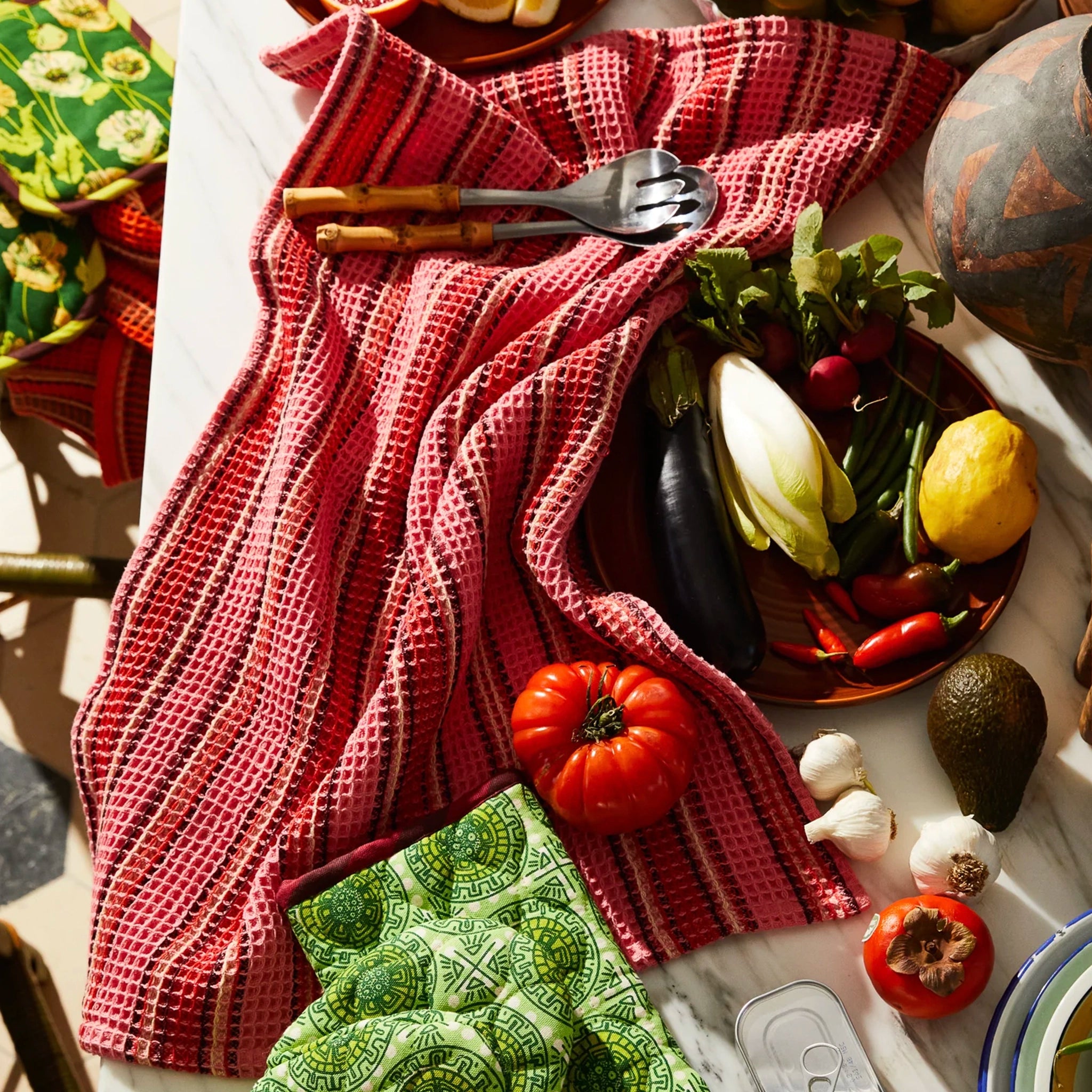 Table setting with a red striped tea towel, green patterned oven mitts, and various fruits and vegetables.