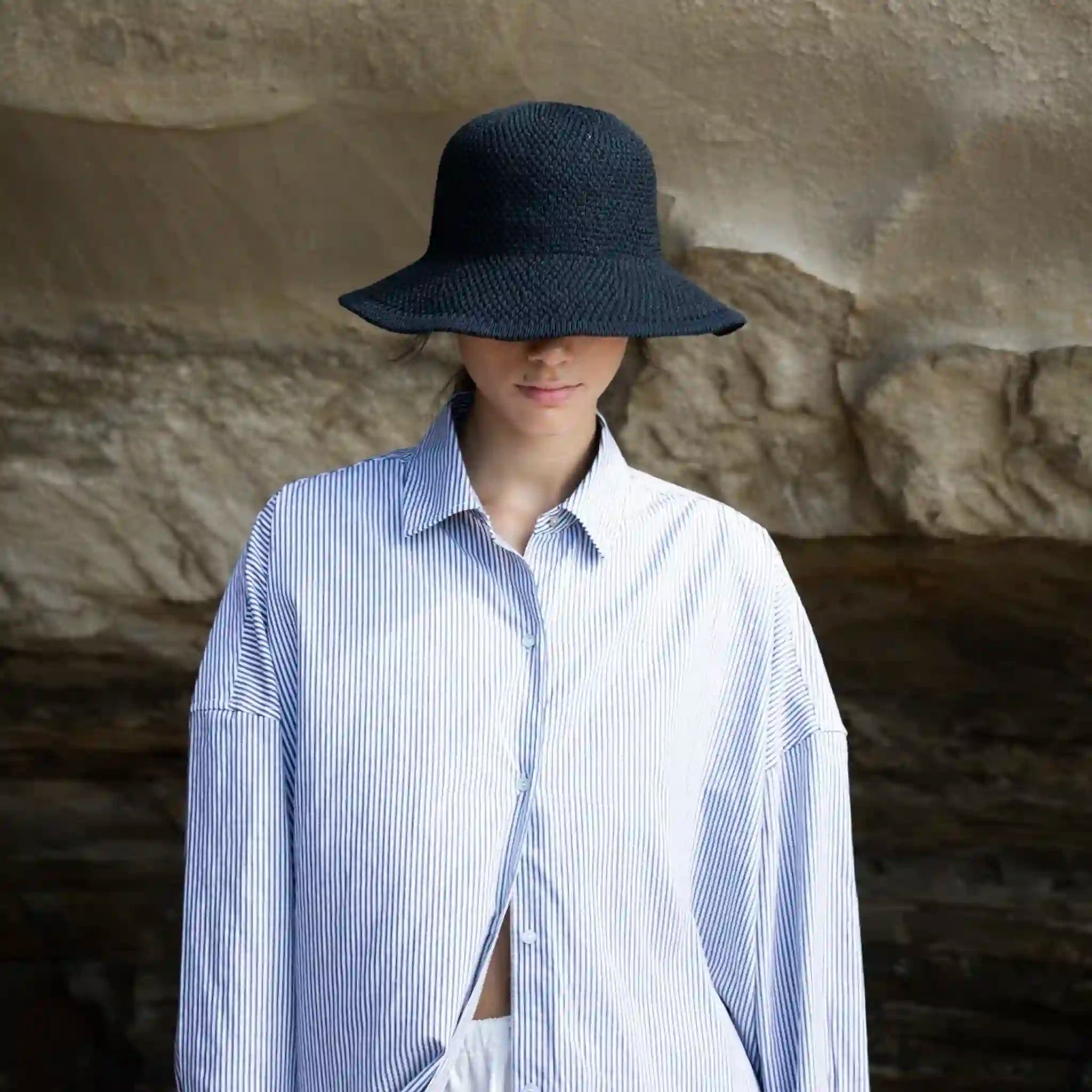 Person wearing a black hat and blue striped shirt against a stone wall.