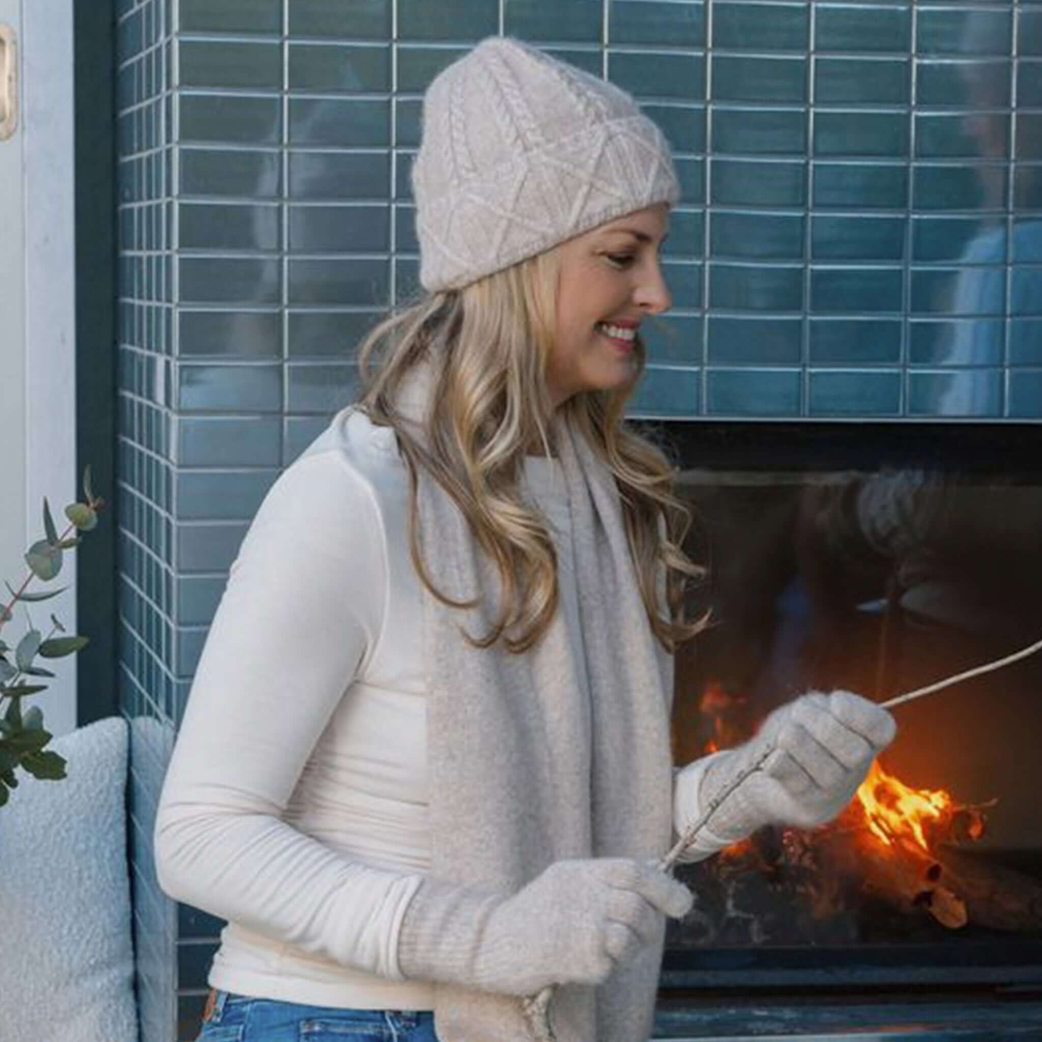 Woman wearing a Sundaise Hotham Cashmere & Wool Beanie - Light Oat, enjoying a cozy moment by the fire.