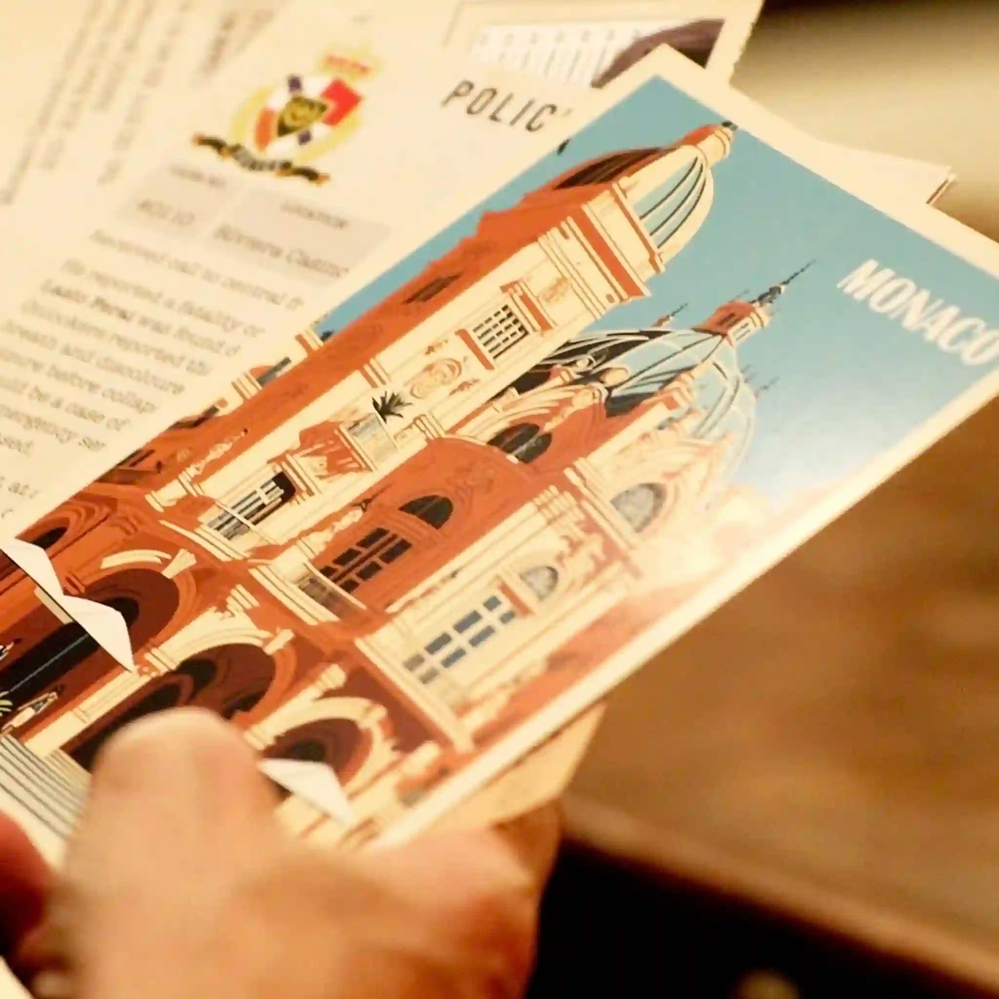 Person holding a postcard with a building illustration and 'Monaco' text, blurred background