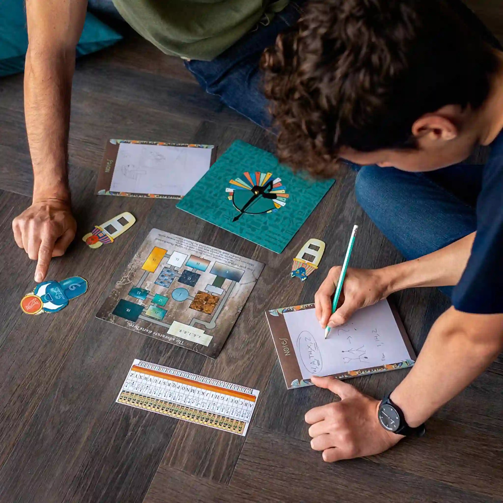 Two people playing a board game on a wooden floor with various game pieces and cards.