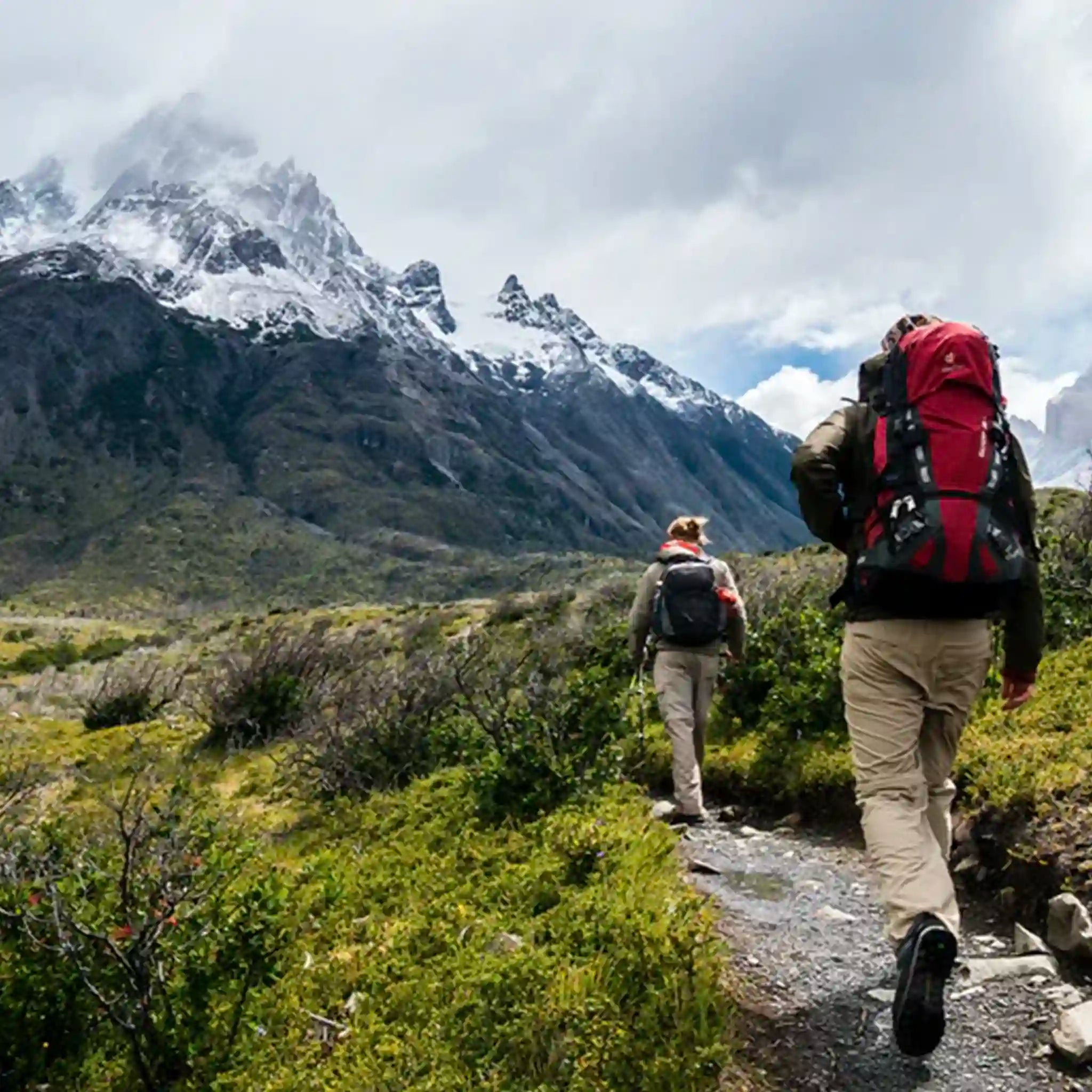 Two hikers with backpacks walking on a trail with snow-capped mountains in the background.