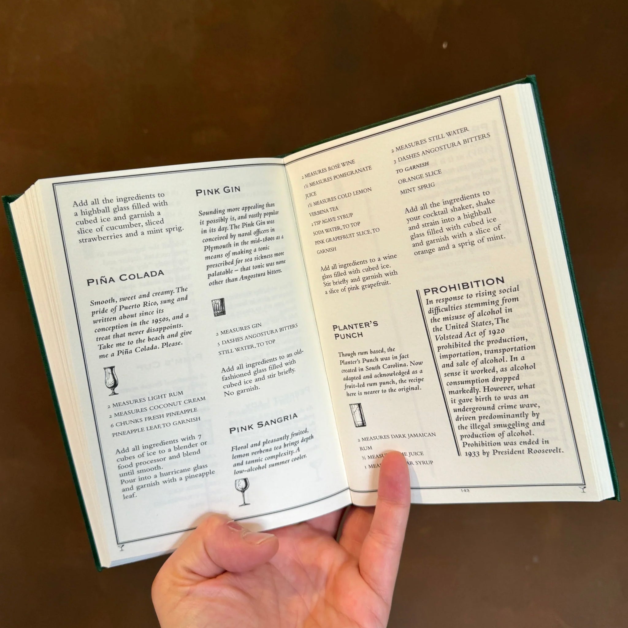 The Cocktail Bible open to pages featuring Piña Colada, Pink Gin, and Prohibition cocktail recipes.