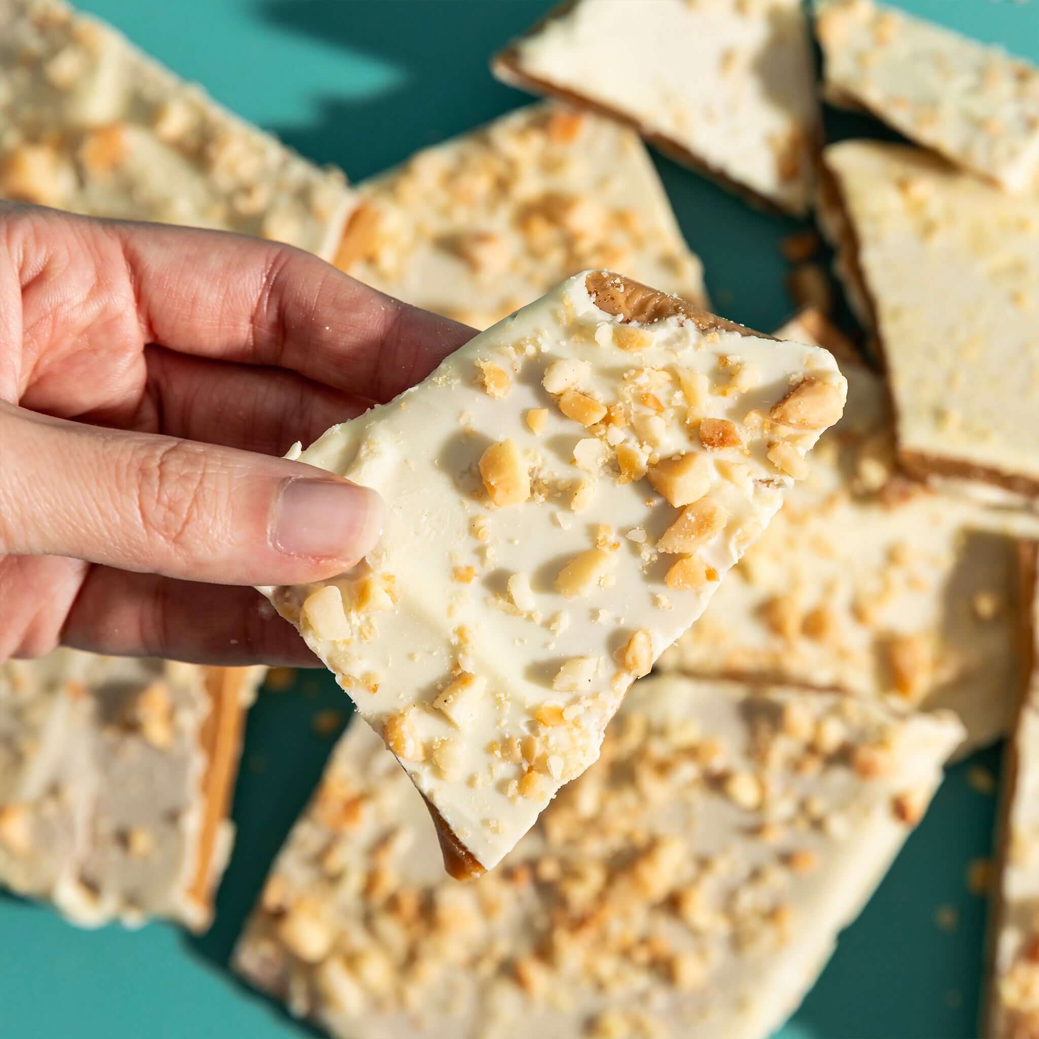 The Confectionist Limited Edition White Chocolate, Macadamia & Lemon Toffee - Large piece held above multiple bark pieces.
