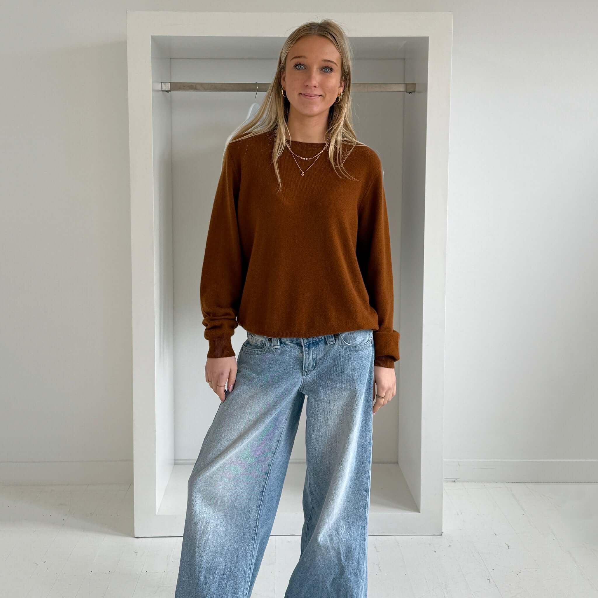 The Uniform Cashmere Crew Neck Sweater - Saddle worn with light blue jeans in a chic indoor setting.