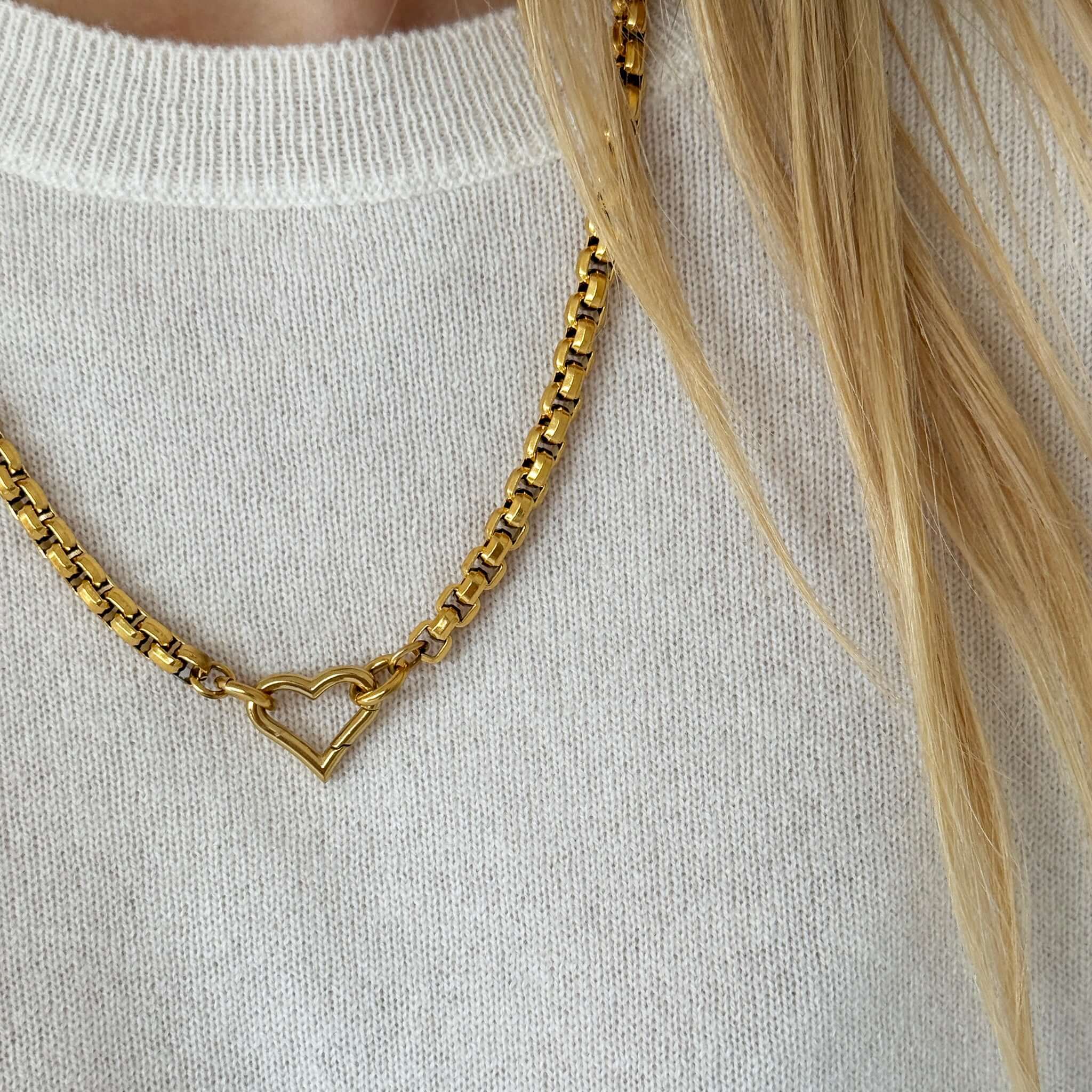 Close-up of a gold chain necklace with a heart pendant, worn over a soft cream sweater.