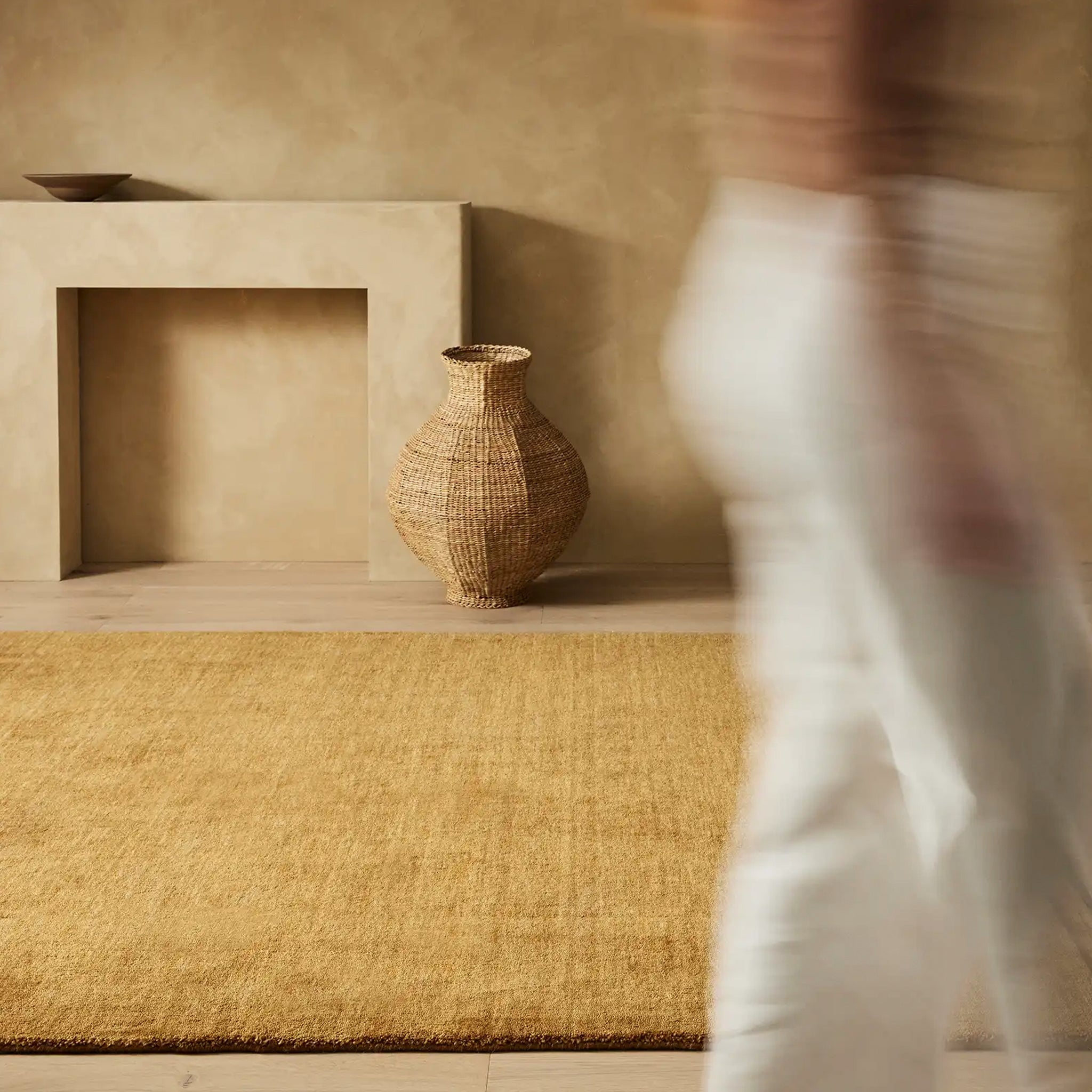 A contemporary rug in a beige color, featuring a textured design and a fringe edge, placed on a floor next to a wall with a vase and fireplace.