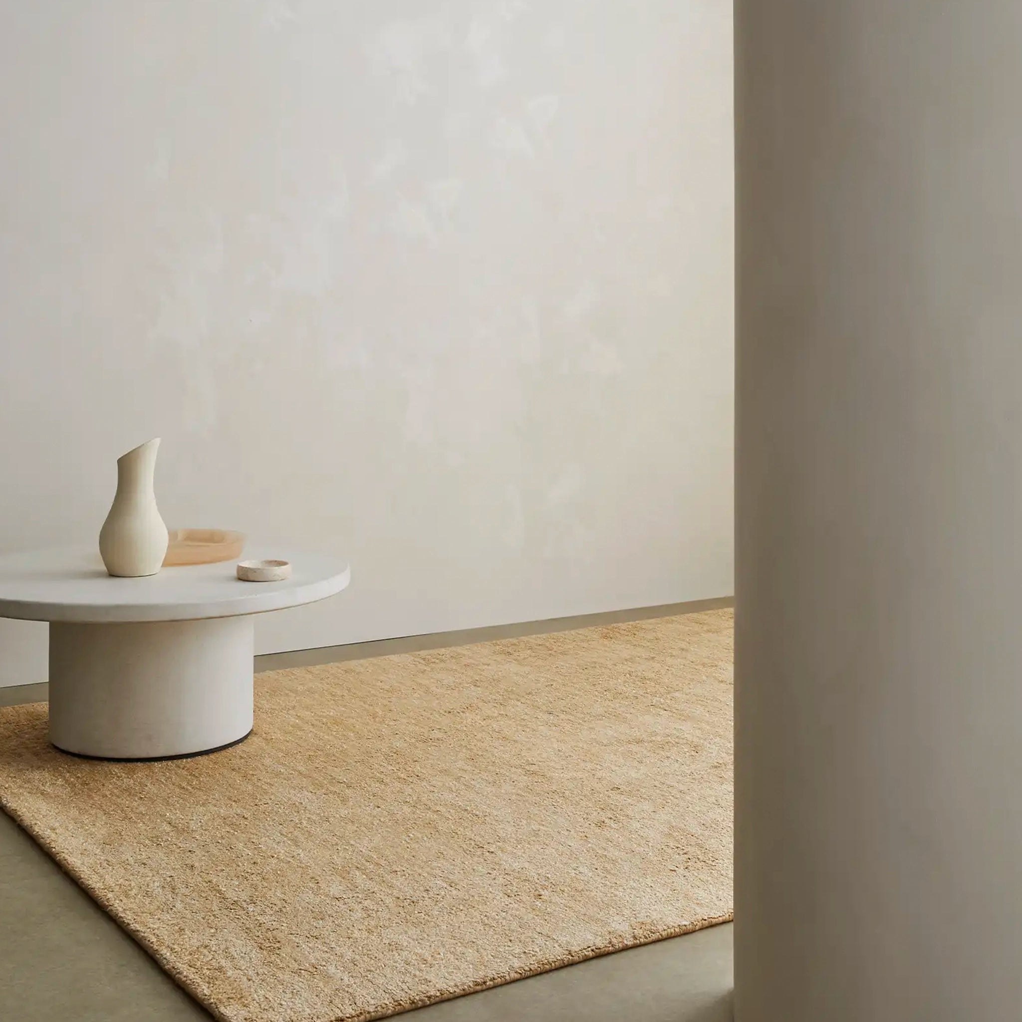 Beige rug on a neutral-colored floor with a white wall and a small round table with a vase.