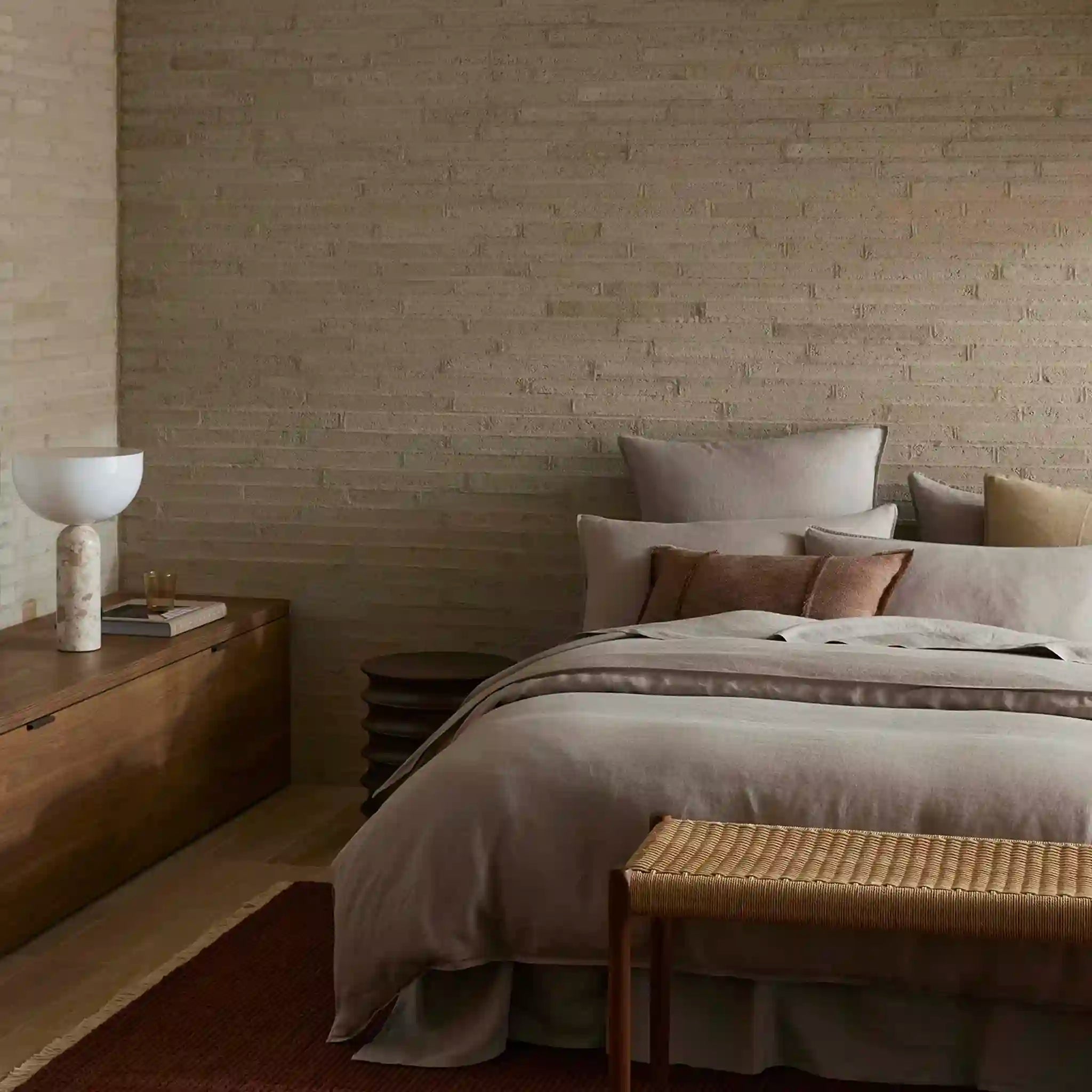 Bedroom with wooden dresser, bed with pillows, and woven bench against a brick wall.