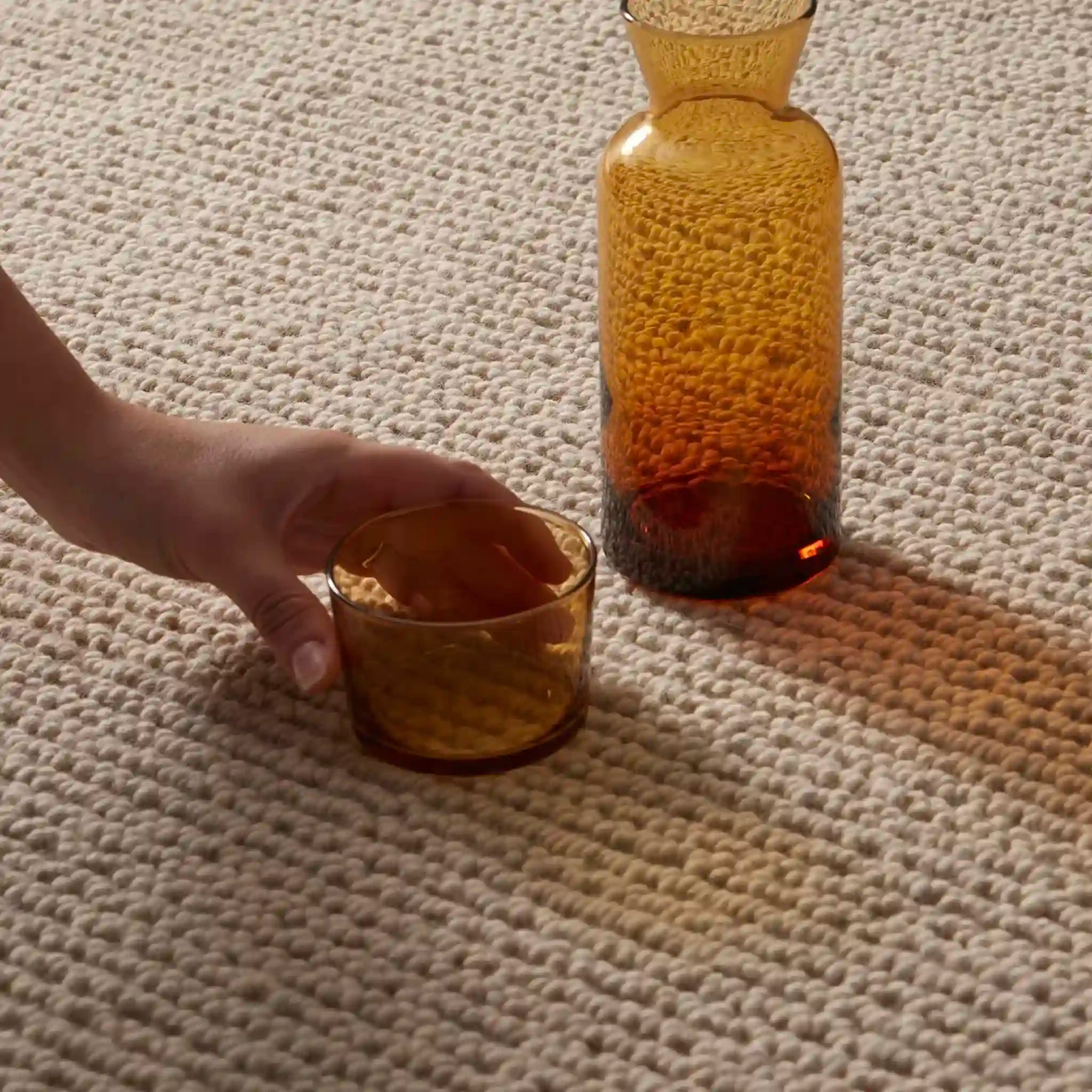 Hand holding a small amber glass container next to a larger amber bottle on a textured surface.
