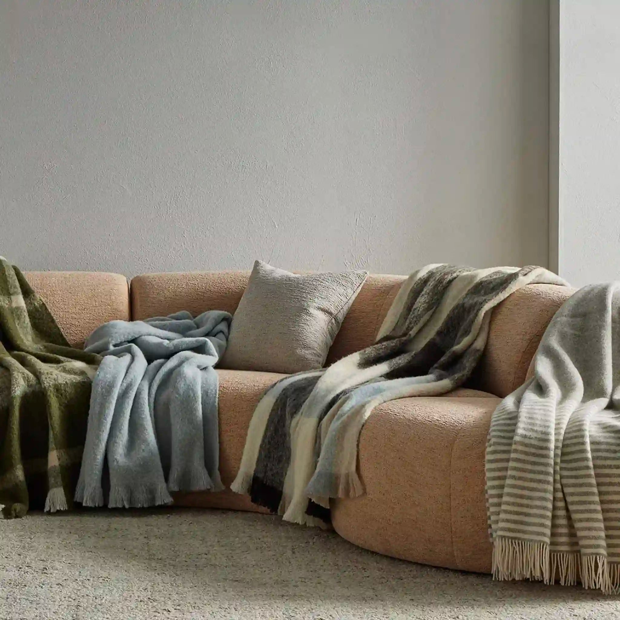 Beige sofa with multiple blankets and pillows against a plain wall.