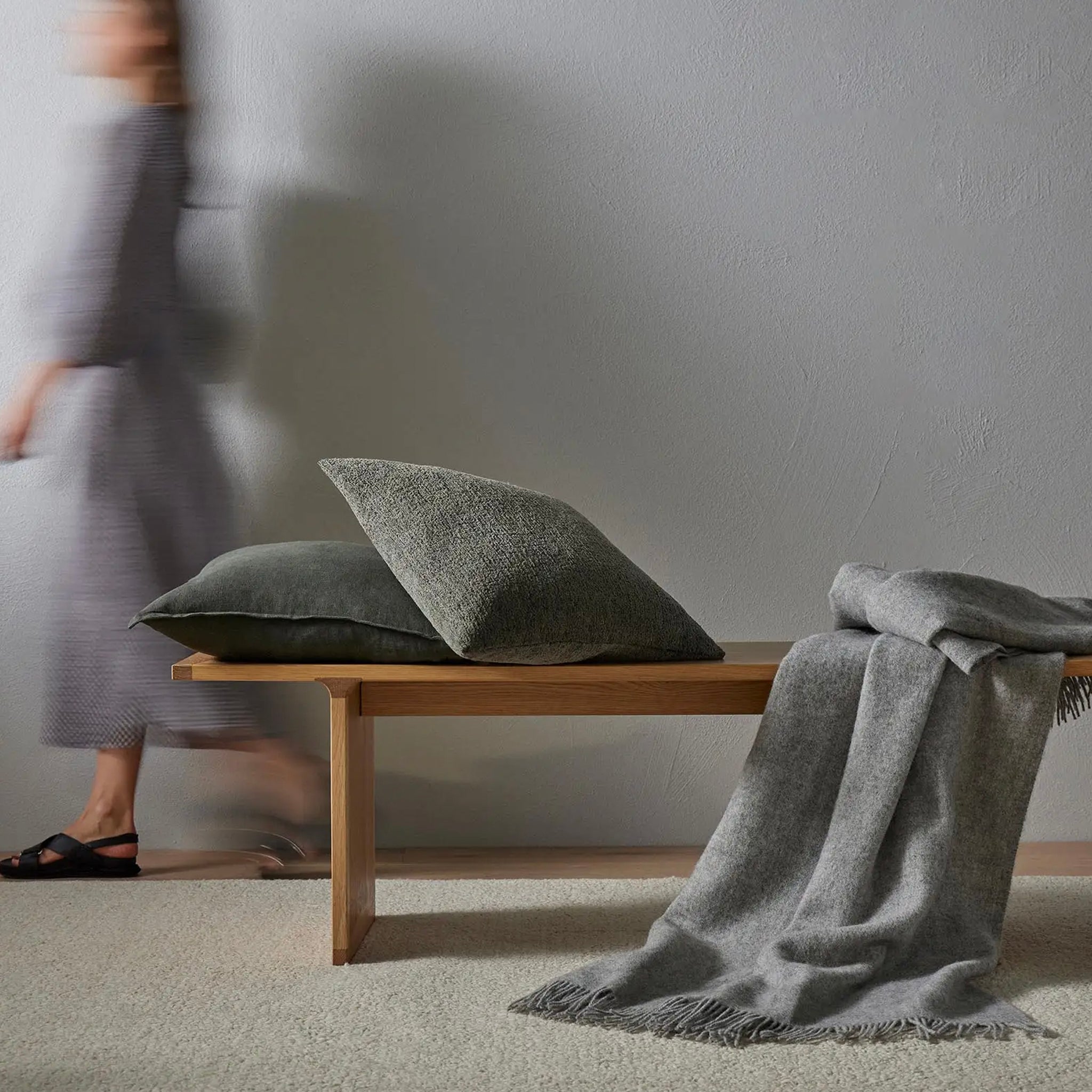 Gray cushions and blanket on a wooden bench with a blurred person in a gray dress walking by.