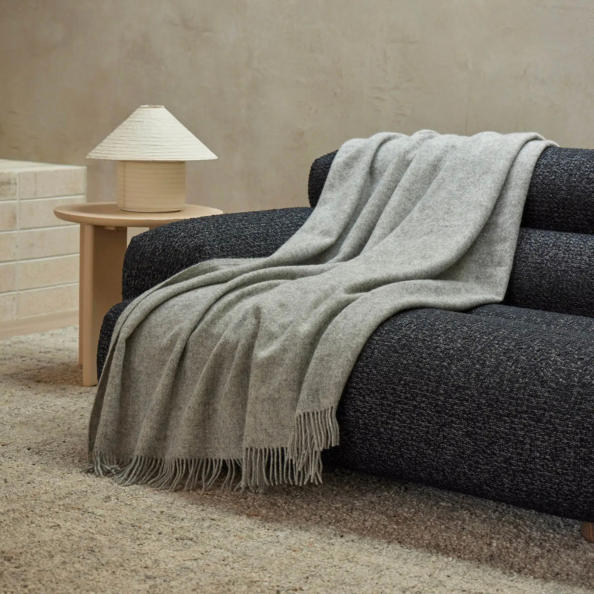 Gray blanket draped over a dark gray sofa with a wooden side table and lamp in the background.