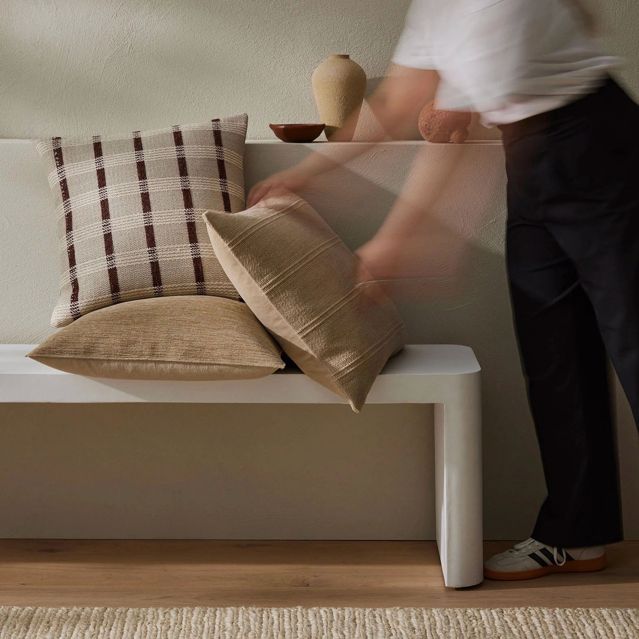 Person arranging pillows on a white bench in a room with a neutral color scheme.